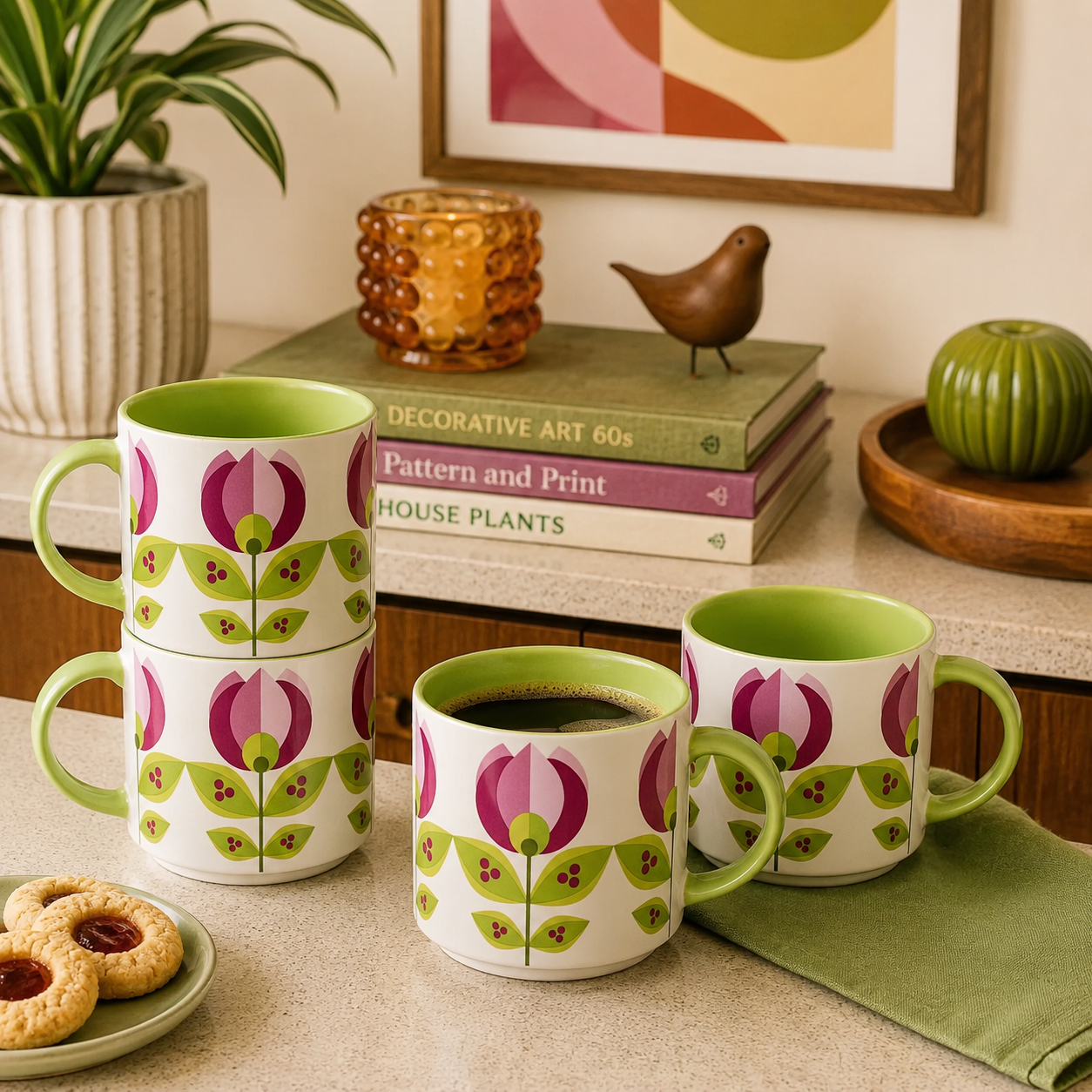 Four ceramic mugs with a white exterior, green handles and interiors, and a repeating tulip pattern in pink and green, arranged on a kitchen counter with books, a plant, cookies, and decorative objects in the background.